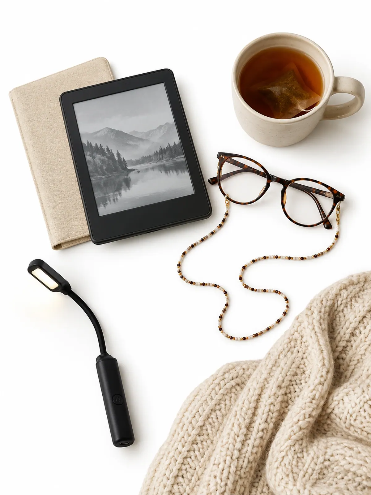 Practical Christmas gifts for women over 50: an e-reader in a cream cover, tortoiseshell reading glasses on a beaded chain, a book light and a mug of tea on a knitted throw