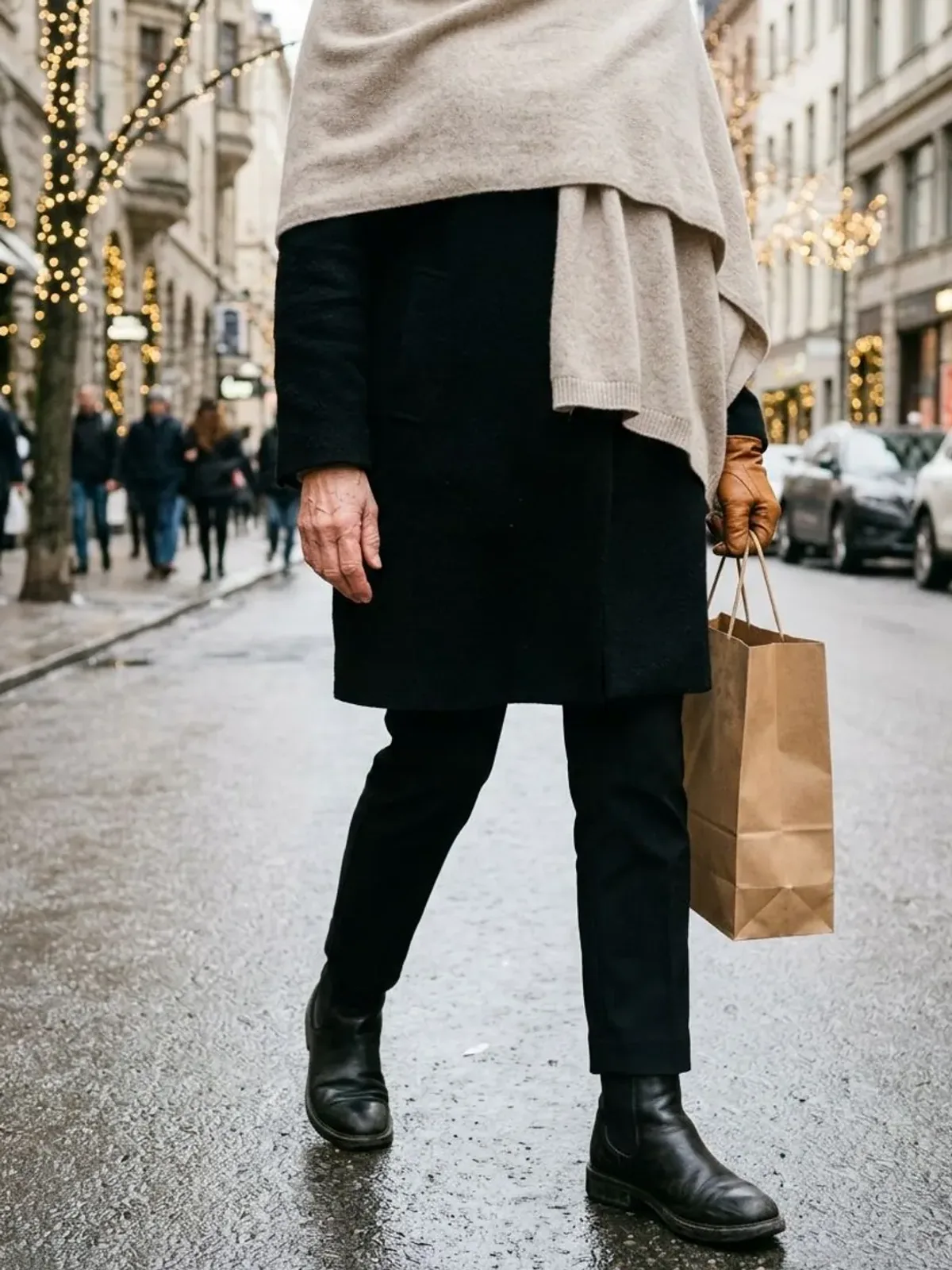 Neck-down view of a woman in her early 60s on a winter street wearing an oatmeal cashmere blanket wrap over a black coat with tan leather gloves and a paper shopping bag