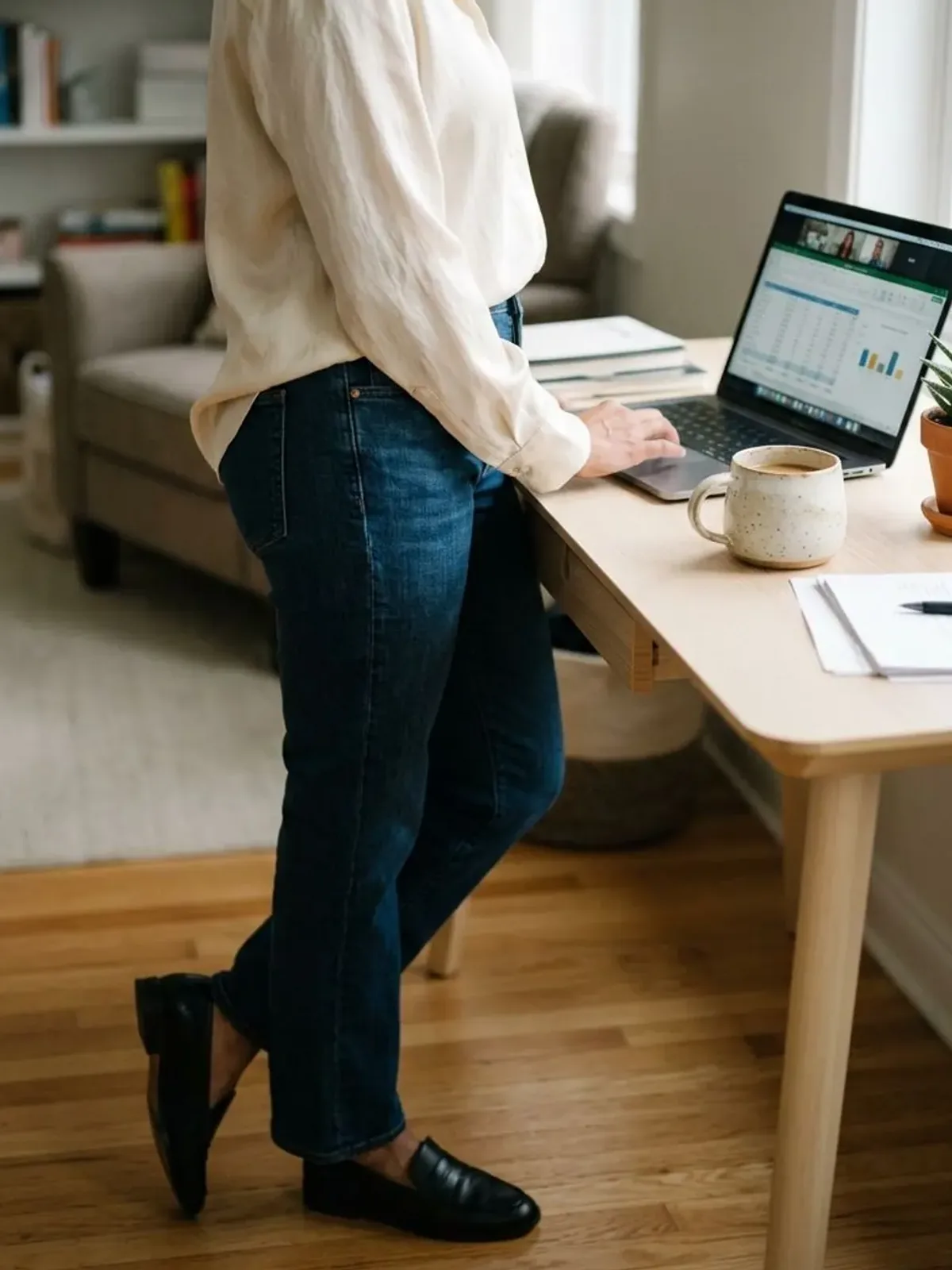 Neck-down view of a woman in her late 40s at a home office desk in a cream satin blouse tucked into dark straight jeans beside a laptop