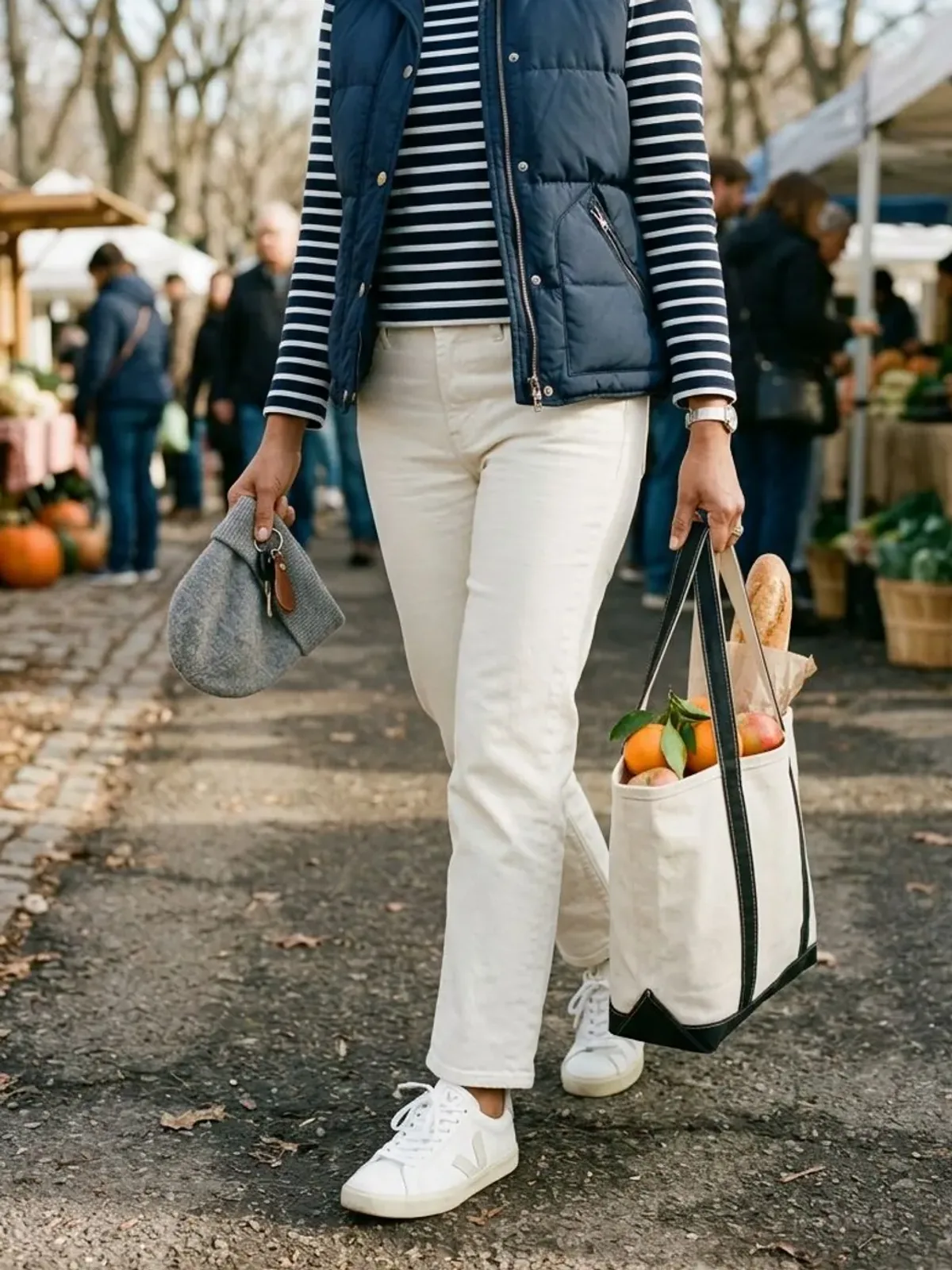 Neck-down view of a woman in her late 40s at a winter farmers market in a navy quilted vest over a Breton striped top, ecru jeans and white sneakers, carrying a canvas tote