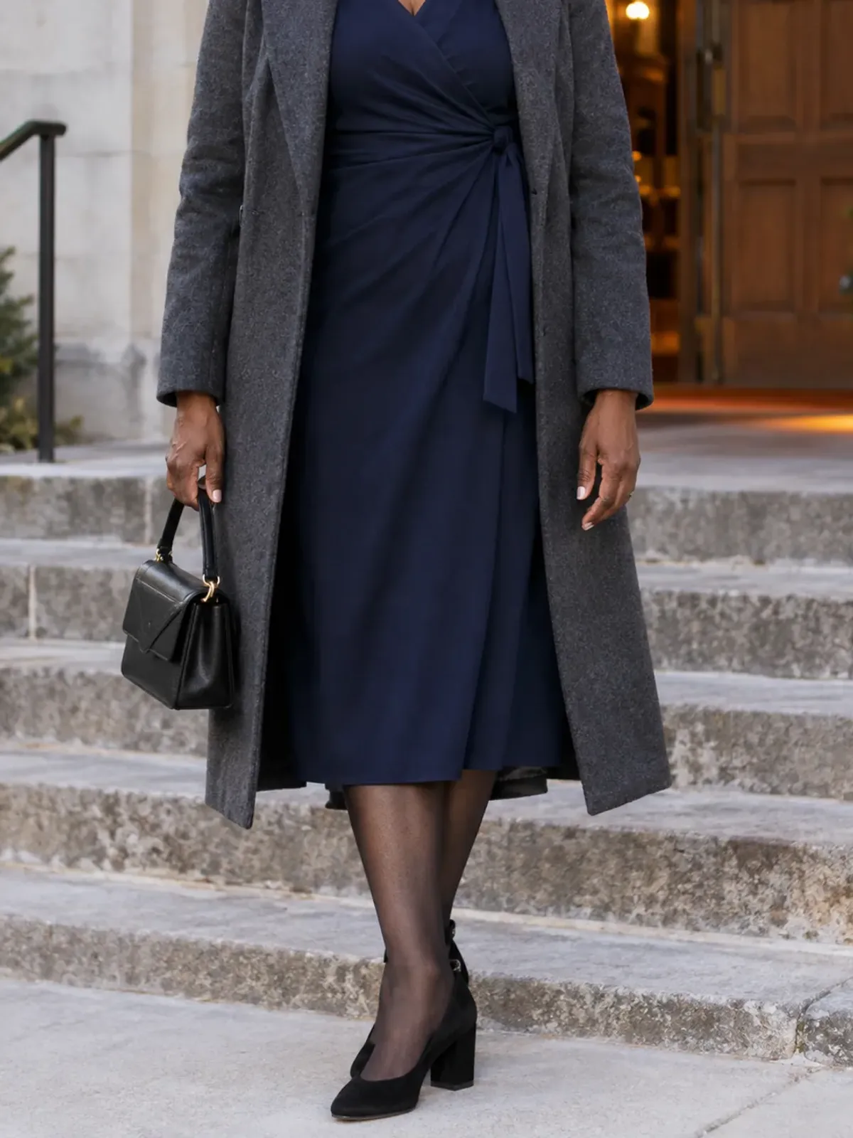 Neck-down view of a woman in her late 50s on church steps at dusk in a long-sleeve navy wrap dress, sheer black tights, black block-heel pumps and an open charcoal coat