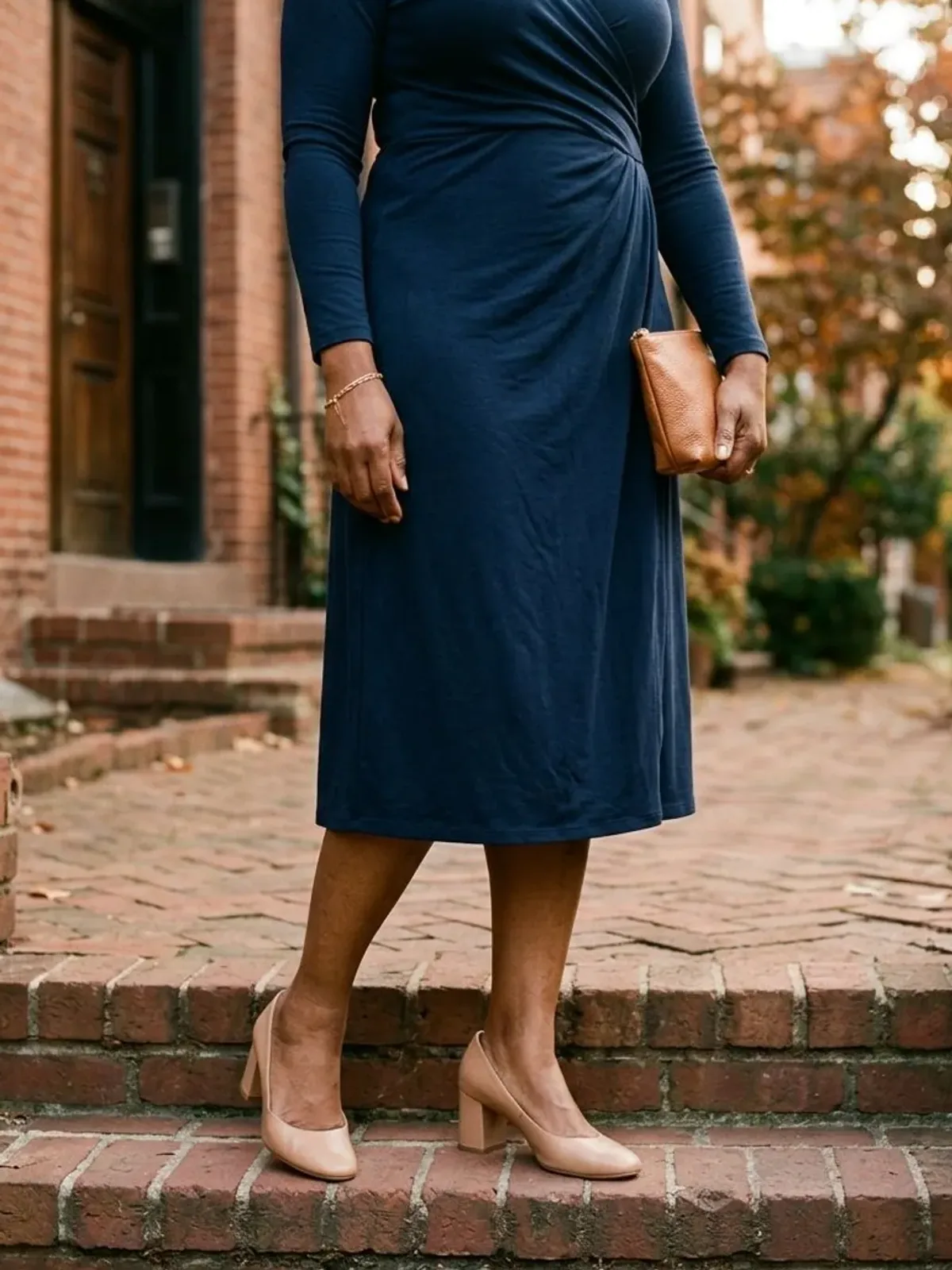 Neck-down view of a woman in her mid 50s on townhouse steps in a navy long-sleeve jersey wrap midi dress with nude block-heel pumps and a small tan clutch