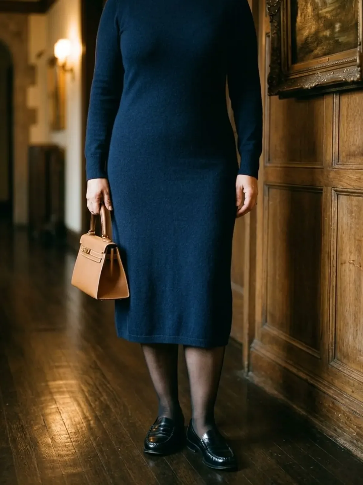 Neck-down view of a woman in her early 50s in a wood-panelled hallway wearing a navy fine-knit midi dress with sheer black tights, black loafers and a small tan handbag
