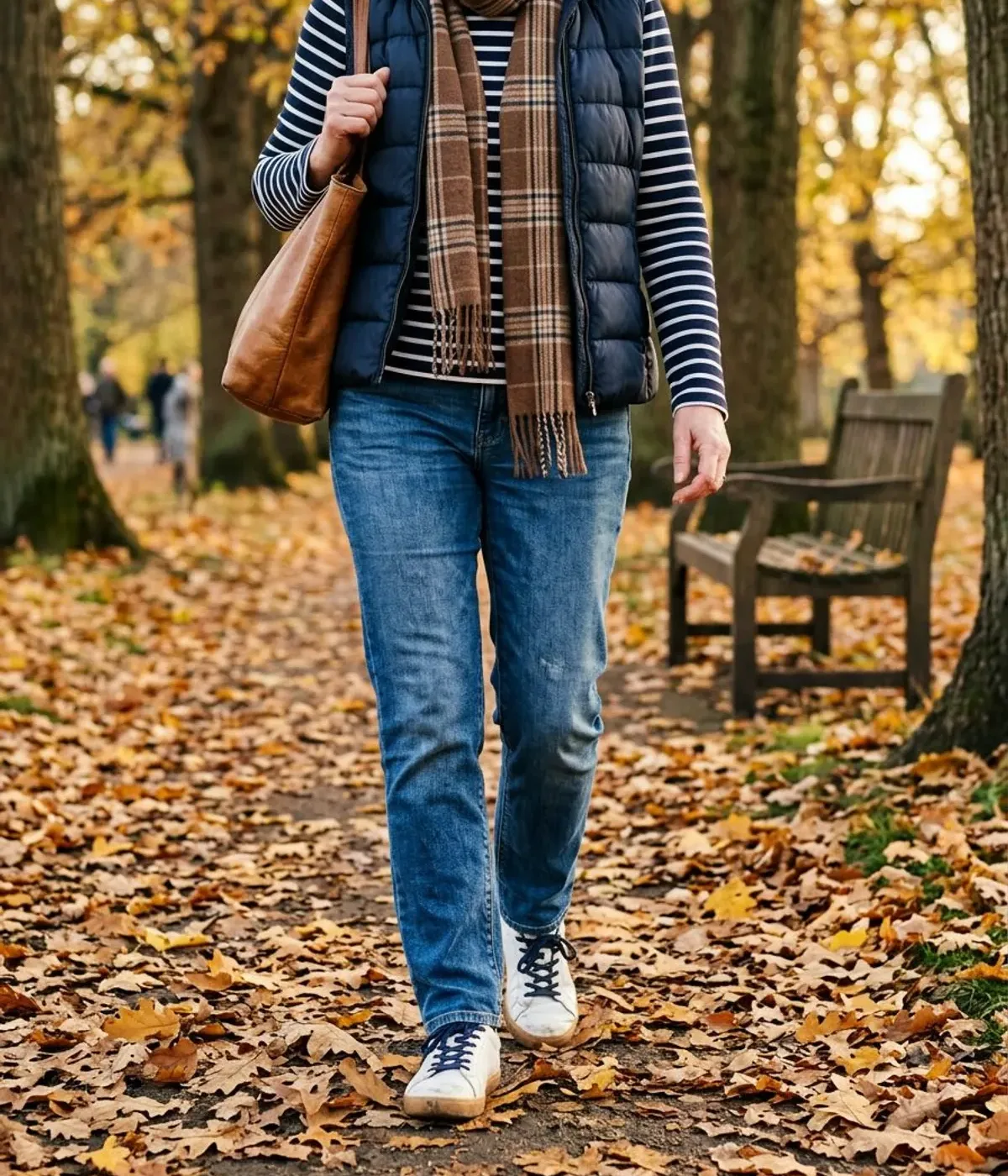 Neck-down view of a woman in her 50s on a leafy park path in a navy quilted vest, striped Breton top, medium-wash straight jeans and white sneakers