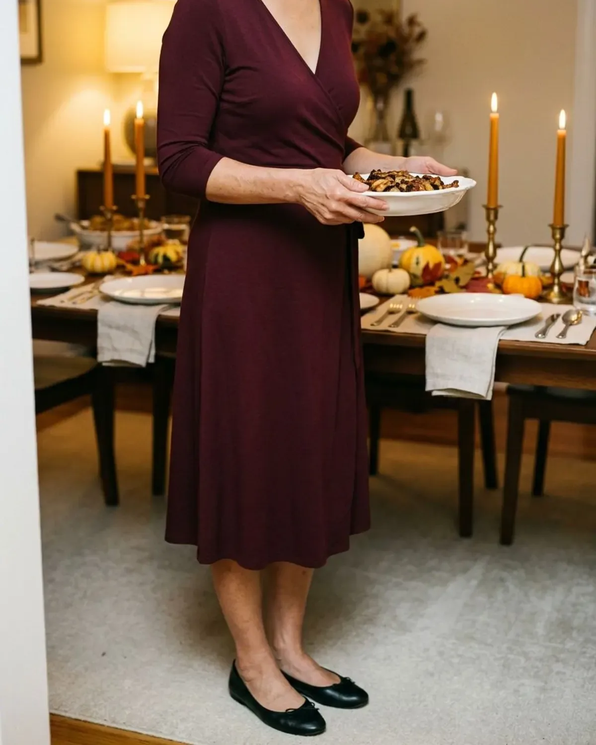 Neck-down view of a woman hosting Thanksgiving in a wine jersey wrap midi dress, black ballet flats and gold drop earrings beside a candlelit table